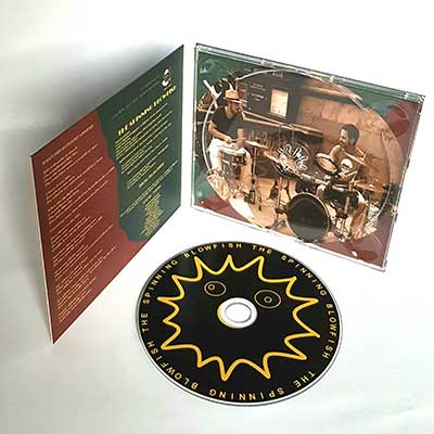 The Spinning Blowfish - Digipak Edinburgh 20 A CD case with booklet features an image of two artists on drums inside, and a CD labeled "The Spinning Blowfish" with a sun-like design—capturing the energy of live music.
