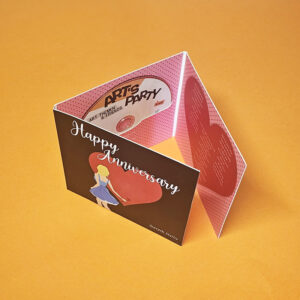 A folded "Happy Anniversary" greeting card with an illustration of a woman holding a heart, displayed on an orange surface. The inside features a pink design, a party invitation insert, and is inspired by Edinburgh’s vibrant celebration style.